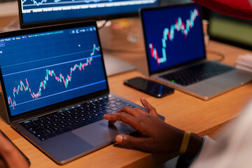 A person's hands are shown interacting with a laptop displaying stock market charts. Multiple screens with financial data are visible in the background.