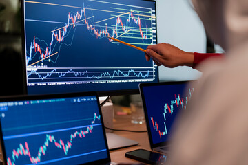 A person points at a stock market chart on a computer screen, analyzing financial data with multiple monitors displaying trading graphs.