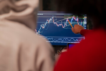 Two people analyze a financial chart on a computer screen, one pointing with a pencil to a specific point on the graph.