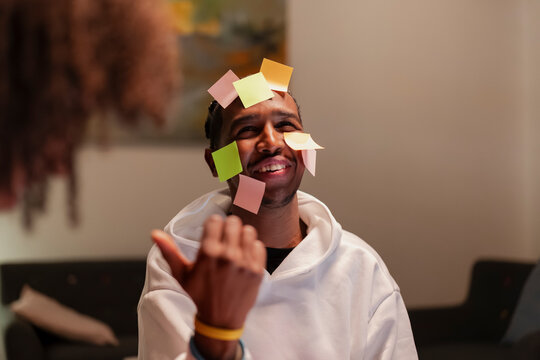 A man with colorful sticky notes on his face smiles and gestures while playing a game with someone out of frame.