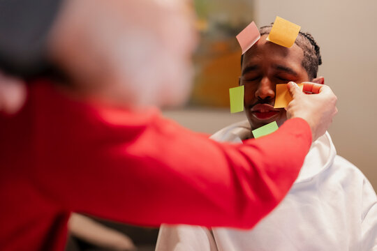 A person in a red shirt places colorful sticky notes on the face of another person wearing a white hoodie.