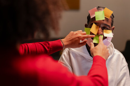 A person places colorful sticky notes on another person's face, creating a playful and humorous moment of connection and lighthearted interaction.