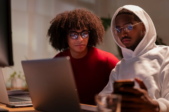 Two young men collaborate on a project, one using a laptop and the other checking his phone, both focused on their tasks in a dimly lit room.