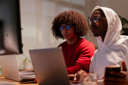 Two young men collaborate on a project, focused on their laptops and a mobile device in a modern office setting.