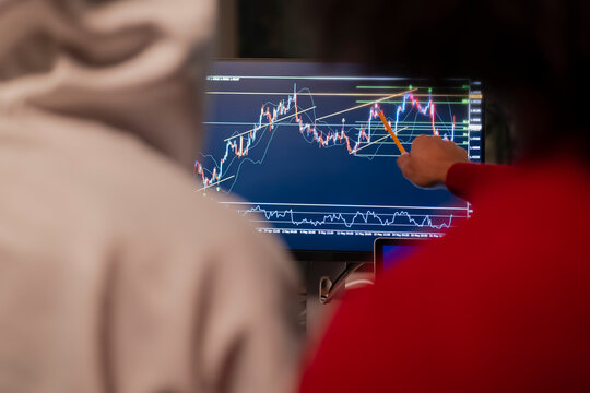 Two people analyze a financial chart on a computer screen, one pointing with a pencil to a specific point on the graph.