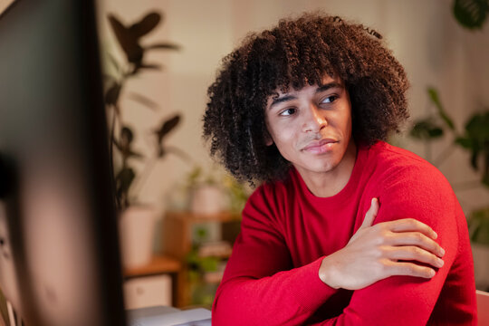 A young man with curly hair wearing a red sweater sits at a desk, looking thoughtfully to the side.
