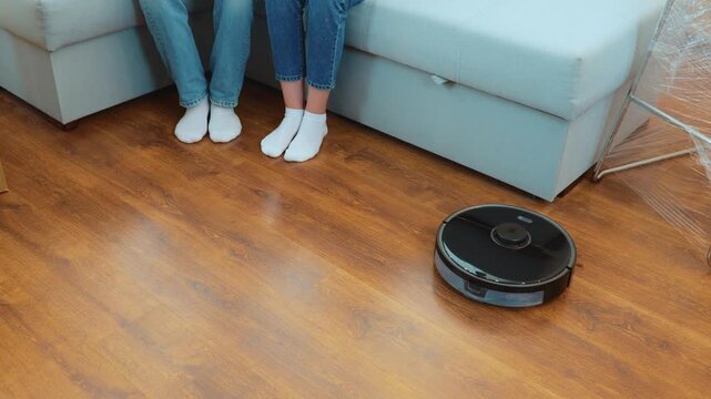 Modern vacuum robot cleaning at home. Close up. Cleaner vacuuming parquet in room. Couple raising their legs while sitting on couch. High angle view. Cleaning robot. Technology concept