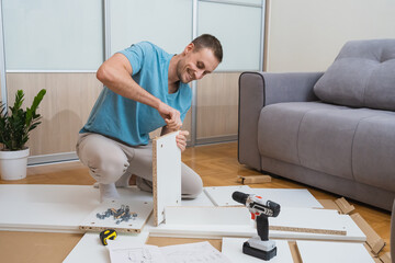 smiling man in a blue t-shirt kneels on the floor assembling furniture parts with a screwdriver and instructions in a bright room