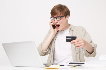 Studio shot of young man sits at a table, holds a credit card in his hand, talking to a bank manager, looks at the laptop screen with happiness, payment operation was successful