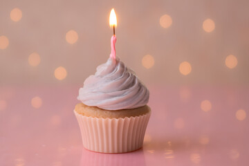 A cupcake with a lit pink candle on top of swirled purple frosting, sitting on a pink surface with a blurred background of warm lights
