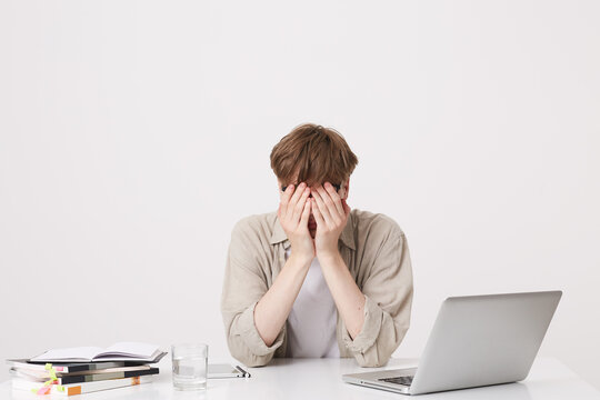 Closeup of tired desperate young man student wears beige shirt coveres face by hands sitting at the table with laptop computer and notebooks isolated over white background