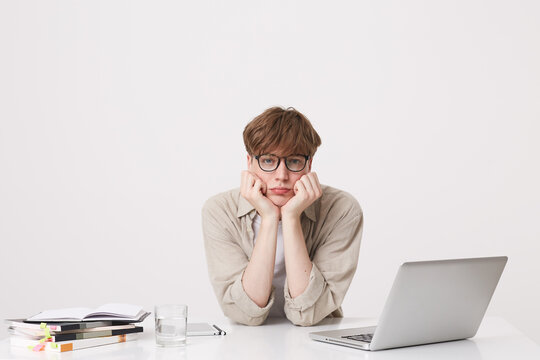 Portrait of upset unhappy young man student wears beige shirt and glasses feels sad and study at the table with laptop computer and notebooks isolated over white background