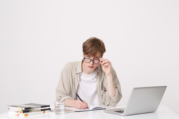 Portrait of concentrated young man student wears spectacles and beige shirt writing and study at the table with laptop computer and notebooks isolated over white background
