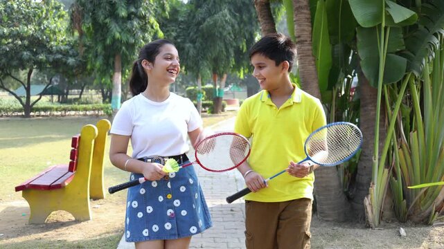 Happy Indian kids walking together in park, carrying badminton rackets enjoying outdoor sport activity. childhood fun. Brother sister talking to each other while walking. Summer Vacations.