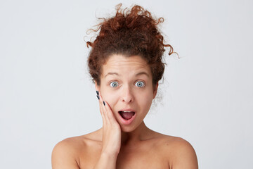 Cropped shot of impressed astonished young woman with curly hair, perfect healthy skin and opened mouth touching face by hand after applying mask or cream and looks amazed isolated over white wall