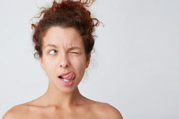 Close up shot of thoughtful playful young woman with curly hair and perfect skin shows tongue, winks and looks to the side isolated over white background Thinking about her job before taking shower