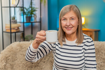 Caucasian mature woman sits on sofa with a cup of coffee smiling as raises cup in greeting or gratitude cheers gesture at home. Grandmother drinking hot drink in the morning, enjoying relaxing resting