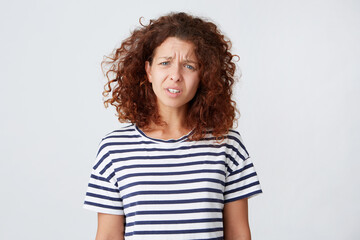 Portrait of sad stressed young woman with long curly hair wears striped t shirt feels irritated and arguing isolated over white background Looks directly in camera