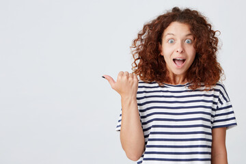 Closeup of amazed cute curly young woman with opened mouth wears striped t shirt looks surprised and points to the side with finger isolated over white background