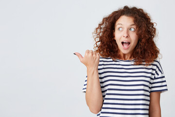 Portrait of surprised beautiful young woman with curly hair and opened mouth wears striped t shirt looks and points to the side isolated over white background