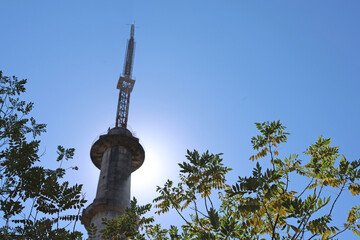 Mhaismal, MH, India, Dec 12: Low angle view of the base of the concrete Doordarshan tower, silhouetted by the sun and framed by dark tree branches and foliage against a bright sky.