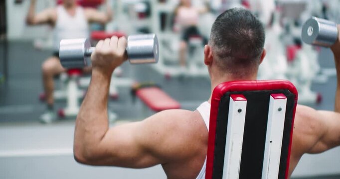 Seated bench press with dumbbells for the deltoid muscles. Deltoid workout with dumbbells. Back view of a middle aged brutal Caucasian man during gym workout in front of a mirror. 4k footage.