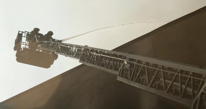 Silhouette of firefighters in a bucket on the ladder of a fire truck, extinguishing with a water gun the roof of a burning building, low-angle view.