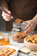 Woman putting tasty granola into glass jar on light grey table, closeup