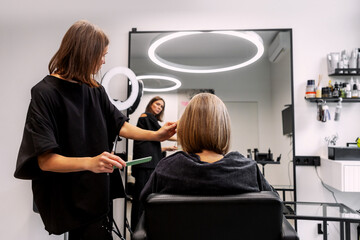 Professional hairstylist shaping neat bob haircut for female client in modern beauty salon, working under bright studio lights inside contemporary interior