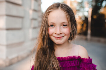 Cute blonde smiling child girl 6-7 year old with long hair and freckles close up outdoors. Looking at camera. Childhood.