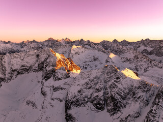 Aerial view of snow-covered mountains during sunrise with golden light reflecting off peaks