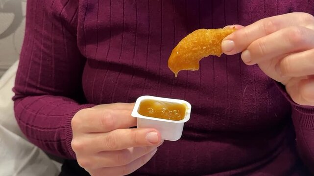 Woman Hand Holding Chicken Nugget Over Sauce, CloseUp Of Casual Eater In Maroon Shirt Dipping Golden Crispy Nugget Into HoneyMustard. Remote Worker Enjoying Quick food.