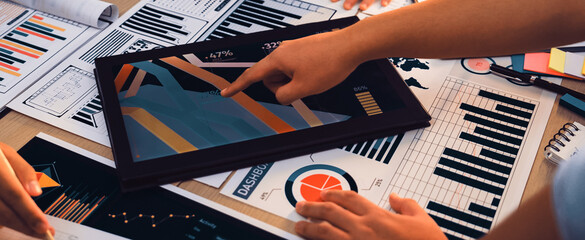 A diverse group of individuals engages in collaborative data analysis using a digital tablet, surrounded by printed charts and graphs in a modern workspace environment. SACTR