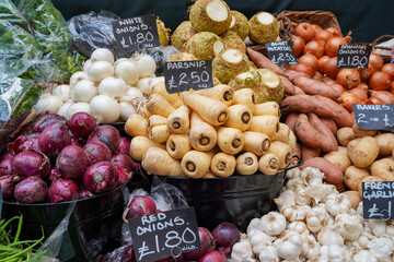Fresh organic root vegetables displayed at local market stall, including parsnips, onions, garlic, sweet potatoes and turnips. Assorted produce arranged in baskets with handwritten price tags.