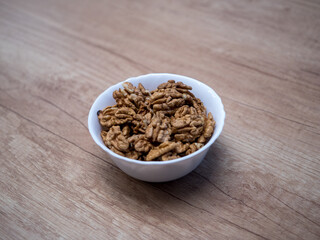 Top view of white bowl filled with raw walnut kernels on wooden table. Natural food ingredient rich in nutrients, suitable for healthy breakfast, baking cakes, desserts and home cooking.