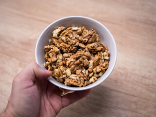Hand holding white bowl filled with raw walnut kernels over wooden background. Natural healthy food rich in nutrients, suitable for baking, cooking, healthy breakfast and snacks.