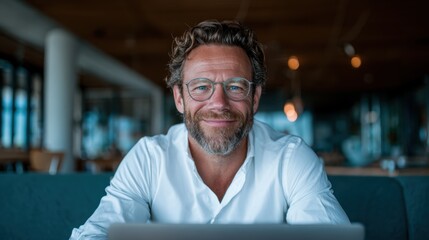 A cheerful man with a beard smiles while working at his laptop in a cozy cafe, capturing the essence of remote work and relaxing environments, ideal for lifestyle stock images.