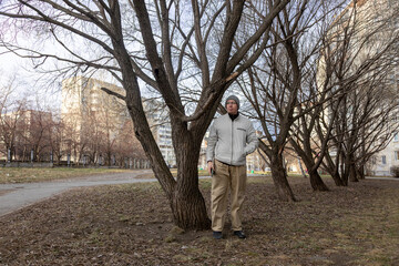 Adult man in city park stands by tree, holding smartphone, waiting for a meeting, looking bored and contemplative.