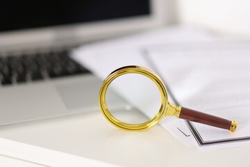 Magnifying glass, laptop and documents on white table, closeup. Space for text