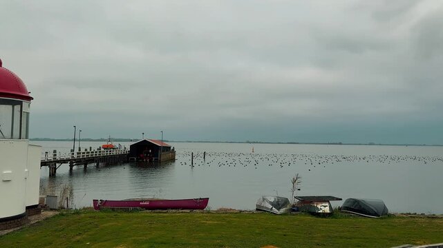 Scenic view of a white lighthouse with a red dome at a Dutch harbor. Features a wooden pier, small boats, and birds on the water under overcast skies.

