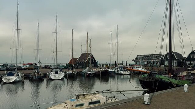 Scenic panning shot of traditional Dutch marina featuring green wooden houses, brick walkway, and various sailboats moored in calm waters under an overcast gray sky in Marken.

