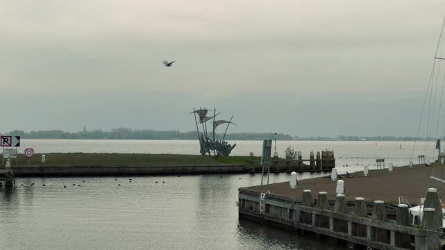 Scenic panning shot of traditional Dutch marina featuring green wooden houses, brick walkway, and various sailboats moored in calm waters under an overcast gray sky in Marken.

