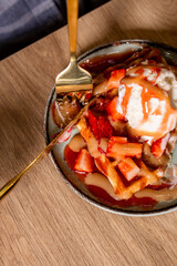 Dynamic dessert moment with a fork cutting into a waffle topped with ice cream, strawberries, and caramel sauce. Warm tones and close-up composition emphasize indulgence and texture.