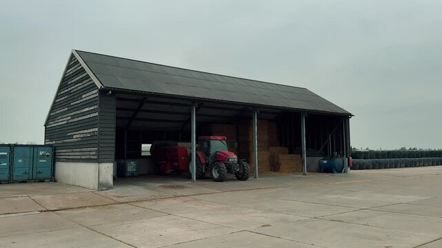 Modern agricultural barn featuring a solar-paneled roof. A red tractor parks inside beside stacked hay, while rows of black wrapped silage bales sit on the concrete outside.

