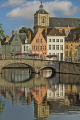 Naklejka premium Canal scene in Bruges featuring a stone bridge with two arches in the foreground and a row of colorful historic buildings with gabled facades along the water.