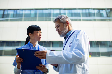 Adult nurse and senior doctor reviewing chart outside hospital, serious