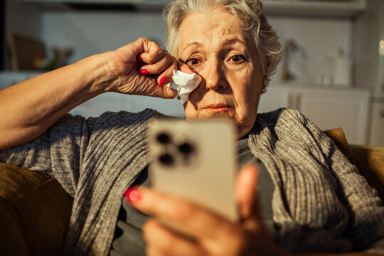 Senior woman sad and wiping tears while looking at smartphone at home