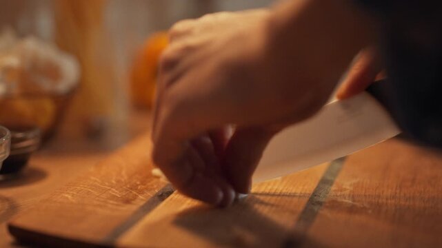 A man's hands meticulously chop fresh garlic cloves with a knife in the kitchen, leveraging its natural spiciness and numerous health benefits for a nourishing evening meal