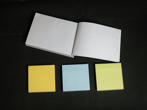 Cuaderno abierto con p&aacute;ginas en blanco y tres blocs de notas adhesivas de colores sobre fondo oscuro