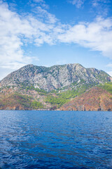 A serene coastal seascape featuring a rocky mountain island covered with patches of green forest rising above a calm deep-blue sea. The clear horizon and bright sky with scattered white clouds